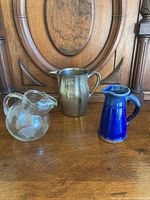 Three pitchers side by side on wooden surface: glass, silver plate, blue pottery