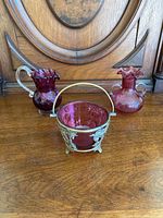 Three vintage cranberry glass items placed on wooden surface against wood furniture; two vases and one bowl with brass casing and handle.