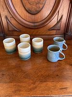 Photo showing four pottery cups with white to greenish-blue gradient glaze and two blue-gray crackled glaze mugs arranged on wooden surface.