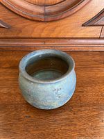 Front view of a small rounded Raku pottery pot sitting on a wooden surface, showing textured rough surface with muted green and blue hues.