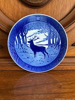 Front view of the 1960 Royal Copenhagen Christmas plate depicting a stag in forest at evening under a star. Plate is blue and white porcelain with intricate border design and embossed date.