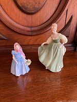 Two Royal Doulton porcelain figurines, 'Fair Lady' in pale green dress and 'Wendy' in blue dress and pink bonnet, displayed on wooden surface.