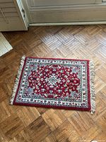 Top view of small red rectangular rug with cream, blue and black floral patterns and white fringes, placed on wooden parquet