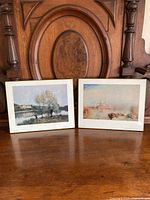 Both mounted art prints propped against carved wooden furniture showing full view of prints and boards.