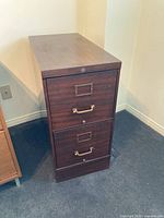 Front angle view showing two drawers, brass handles and label holders, with faux wood grain finish on filing cabinet.