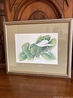 Framed watercolour print of a white lily flower with green leaves, displayed against a wooden background.