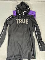 Photo of a black hooded long sleeve shirt with the word TRUE in large letters across the chest, lying on top of various colored tops including purple and dark colors.
