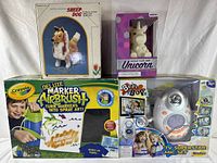 Five toys displayed on white surface: Sheep Dog in box, Design Your Own Unicorn in box, Crayola Deluxe Marker Airbrush set, Bear Bop game, TV Superstar Mic Set
