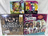 Four boxed items arranged together: Duck Dynasty family party game, Bratz game, Disney Mickey and Minnie 500-piece puzzle, and Disney Fairies 100-piece puzzle.