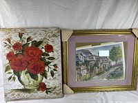 Photo showing both framed artworks side by side: one is a floral painting in a glass vase, the other is a framed lithograph of a house with purple matting.