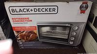 Box front showing Black+Decker branding, rotisserie convection countertop oven image, and capacity detail (holds two 12" pizzas or full-size rotisserie chicken)
