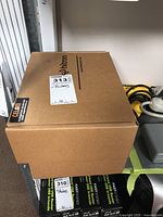 Two Hitron routers in original sealed cardboard packaging, placed on a shelf next to an electrical outlet and other cables.