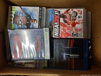 Box of video games including International Cricket 2010 for PS3, NBA 2K11 for Xbox 360, Need for Speed for Xbox 360, and a PlayStation 3 system box.
