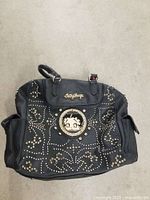 Front view of black Betty Boop satchel handbag showing rhinestone and metal stud embellishments with central Betty Boop character patch and gold-tone Betty Boop nameplate.