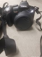 Canon PowerShot SX10 IS digital camera with lens cover and strap, showing front and top body with branding and controls.