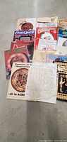 Top view of a spread of 10+ vintage magazines and pattern booklets related to Americana heritage craft handiwork, embroidery, and painting patterns from the 1980s and beyond arranged on floor.