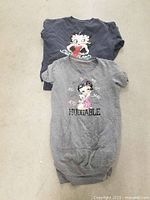Two Betty Boop apparel items laid flat on floor - gray short-sleeve t-shirt dress with "HUGGABLE" Betty Boop print in front and navy long-sleeve sweater with large Betty Boop graphic behind it.