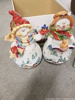 Two Christmas snowman figurines in porcelain style, colorful hats and scarves, set on a surface with a box in background.