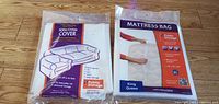 Two new items in sealed packaging laid flat on wood floor: a sofa/chair cover and a mattress bag for king/queen mattresses. Both packages clearly labeled Public Storage.