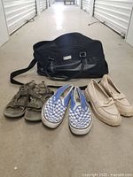 Three pairs of women's shoes placed in front of a large black travel bag. Olive outdoor sandals, blue and white checkerboard Vans slip-ons, white wedge loafers.