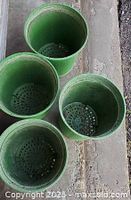 Overhead view of 4 green cylindrical plastic growing pots with drainage holes, showing the interior and the bottom perforations, placed on a cement floor.