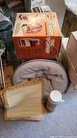 Photo showing boxed West Bend The Poppery popcorn popper, gray padded camp seat, woven straw mat, and thermos container on floor surrounded by boxes and clutter.