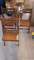 Four wooden folding outdoor chairs arranged in a room with boxes and carpet flooring. Chairs have slatted wooden seats and backs.