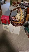 Stack of dark brown woven baskets with the ceramic nativity set on top and green design on pieces. Several cardboard boxes on floor nearby.