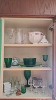 Three-shelf cabinet with various glass and china pieces including white ceramic soup tureen, green glassware, crystal glasses, and small decorative items.