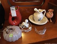 Overview of the lot on wooden surface including glass pumpkin, cherub plug-in light, wash jug with bowl, fish gurglepot, and pink piggy bank