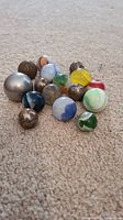 Collection of various marbles placed on carpet, including clear, colored, and metal spheres of different sizes and designs.