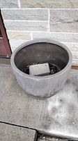 Front view showing interior of composite planter with concrete block and soil debris inside on a concrete surface against a stone wall.