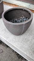 Dark brown composite planter on concrete surface, filled with black decorative rocks and some dry leaves, showing overall condition and size perspective.