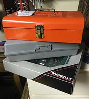 Three stacked toolboxes: an orange metal toolbox on top, a gray plastic case in the middle, and a Mastercut drill bit box on bottom, showing the full lot composition.