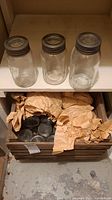 Three vintage glass Mason jars with metal lids on a shelf above a wooden crate filled with wrapped jars
