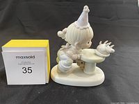Side view of the Precious Moments porcelain figurine showing a child in a party hat blowing out candles on a cake on a pedestal table.
