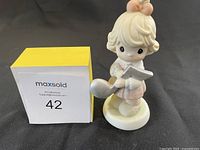 Front view of Precious Moments figurine showing girl holding spoon and musical note, next to Maxsold box numbered 42.