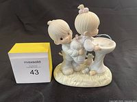Front and side angled view of Precious Moments porcelain figurine depicting two children with bird bath and rabbit, alongside a maxsold lot marker.