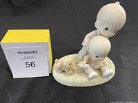 Porcelain Precious Moments figurine with two children and a puppy on an oval base, displayed against a black background with MaxSold auction sign.