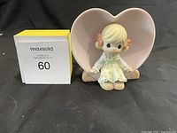 Front view of Precious Moments figurine with a girl in a light blue dress and a large pink heart backdrop.