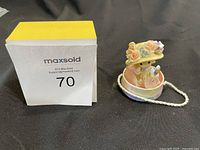 Front angle of the Precious Moments figurine featuring a girl in a hat sitting in a pastel colored hat box with floral accents and white string handle.