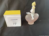 Photo of a Precious Moments figurine depicting a blonde girl in a white dress standing atop a frosted pink glass heart with embossed flowers, next to the MaxSold lot number.
