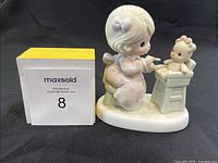 Porcelain Precious Moments figurine showing a young girl pointing at a teddy bear on a chair, with a MaxSold card beside it for scale and ID.