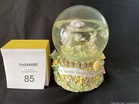 Photo of a Precious Moments music box snow globe figurine titled 'All Things Grow' from 1998. The globe features figurines of a child and a bunny surrounded by flowers, on a floral base with the inscription 'All Things Grow'.
