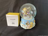 Photo of Precious Moments 1998 Frosty The Snowman music box figurine showing glass globe with two children inside, seated on a blue base decorated with snowflakes, a drum, and a teddy bear figure.