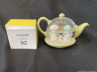 Precious Moments figurine in a teapot glass dome with two children inside, on a yellow base, with MaxSold lot number 92 box nearby.