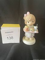 Front view of the Precious Moments figurine showing the girl with a pink bow holding a tray with food items and a gift box, next to a MaxSold lot sign.
