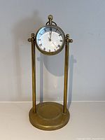 Full front portrait of brass desk clock with circular clock face and orb crystal lens, held by two vertical brass rods on a round base. Shows overall clock design and size.