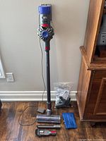 Dyson V7 Motorhead Origin cordless stick vacuum assembled upright with charger plugged in, motorized head attached, accessories laid out