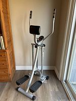 Silver exercise machine with black padded back support and foot pedals, digital display and handle sensors visible